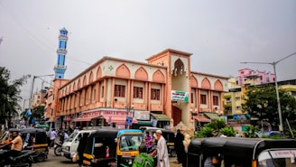 A bustling street scene in an urban area with several autorickshaws and people in the foreground. The prominent building features Islamic architectural elements with arched windows and an adjacent blue and white minaret. The surrounding area includes various shops and residential buildings.
