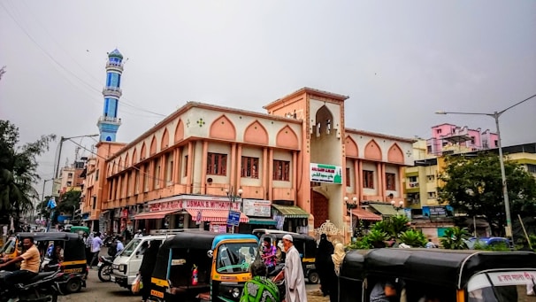 A bustling street scene in an urban area with several autorickshaws and people in the foreground. The prominent building features Islamic architectural elements with arched windows and an adjacent blue and white minaret. The surrounding area includes various shops and residential buildings.
