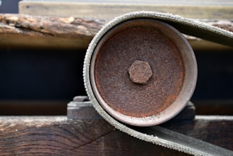 A close-up view of a metal pulley with a textured surface, surrounded by a belt. The pulley has a rusty, corroded appearance, indicating age and wear. The background consists of wooden beams and possibly an industrial setting.