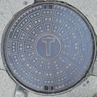 A round manhole cover on a concrete surface. The manhole cover has a geometric pattern with concentric circles and radial lines, filled with small dots. At the center, there is a raised letter 'T'. There are markings around the edge, including 'EN124 D400'. The surrounding surface has slight green moss in some areas.