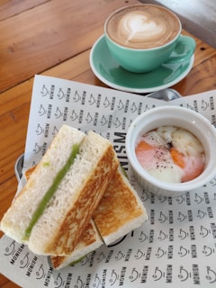 A plate with toasted sandwiches filled with a green spread, a small bowl of soft-boiled eggs with a dash of pepper, and a cup of cappuccino with latte art in a green cup and saucer. The items are placed on a wooden table with a branded paper wrapping underneath.