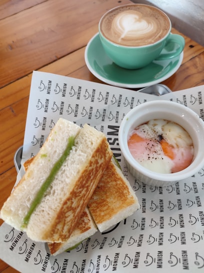 A plate with toasted sandwiches filled with a green spread, a small bowl of soft-boiled eggs with a dash of pepper, and a cup of cappuccino with latte art in a green cup and saucer. The items are placed on a wooden table with a branded paper wrapping underneath.