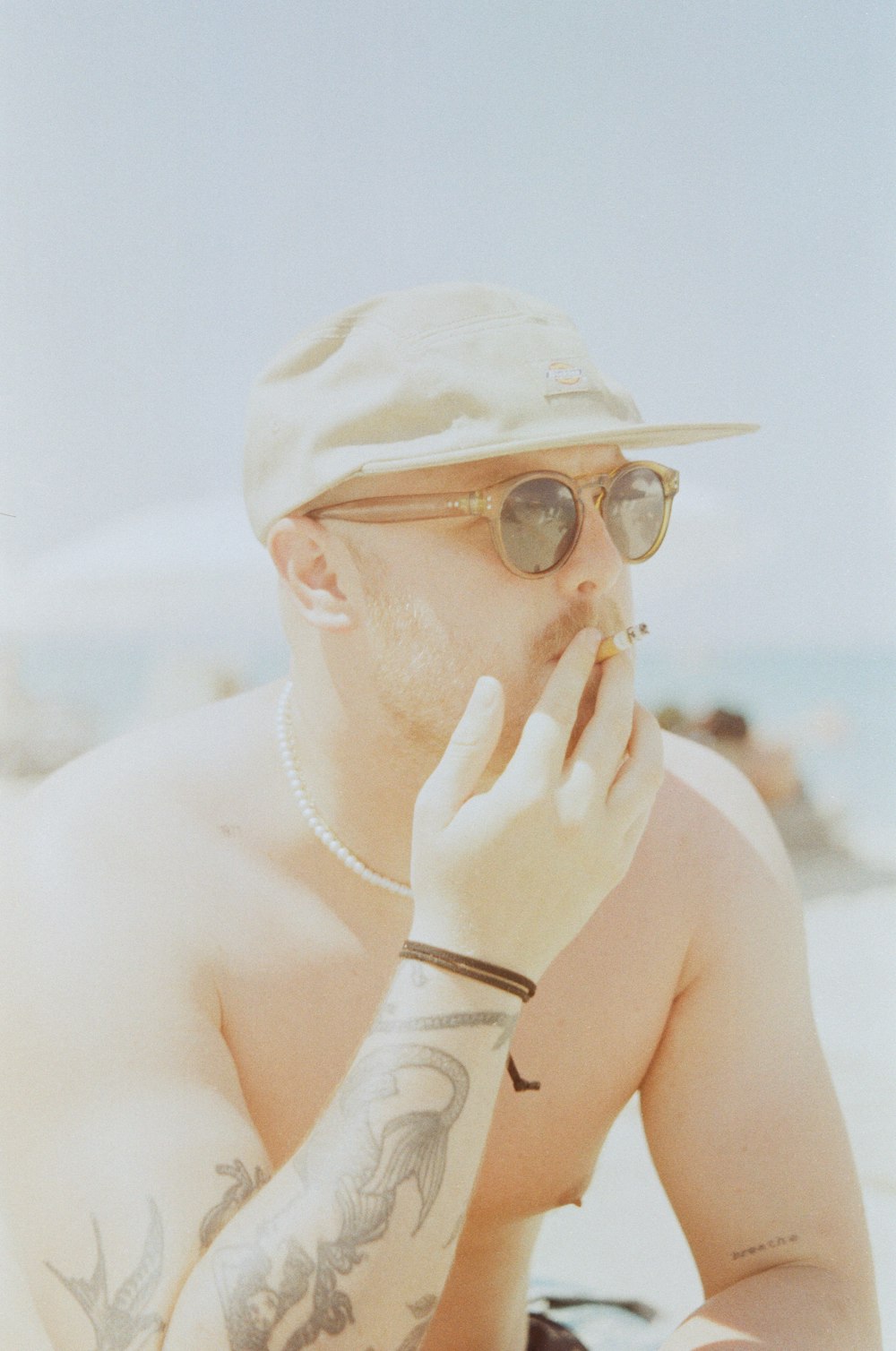 a shirtless man sitting on the beach smoking a cigarette