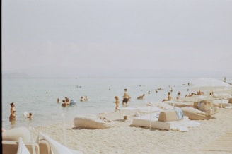 A beach scene with people enjoying the calm, clear water and lounging on the sand. The beach is equipped with cushions and shaded by large umbrellas. The atmosphere feels relaxed and summery.