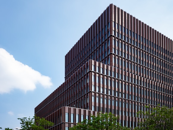 A modern, multi-story office building with a geometric facade made of vertical metal and glass panels. The structure is surrounded by greenery, and the sky is clear with a few clouds.