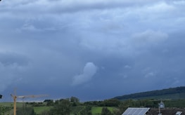 A cloudy sky stretches over a landscape, with dark, thick clouds signaling an impending storm. Below, green fields and trees cover rolling hills. To the left is a construction crane, and on the right, solar panels are visible on a rooftop.