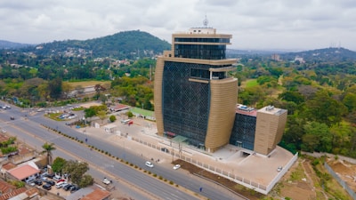 A modern multi-story building with a unique curved design stands prominently along a major road. The structure features a combination of glass and beige panels, giving it a sleek and contemporary look. Surrounding the building are well-manicured green spaces and a parking area. The backdrop includes lush trees and hills, while cars and houses are visible along the road.