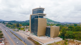 A modern, multi-story building with a unique architectural design stands prominently. It features a combination of glass and concrete materials, with the facade designed in a contemporary style. The surrounding area includes a road with moving vehicles, and lush greenery envelops the scene, with hills visible in the background under a cloudy sky.