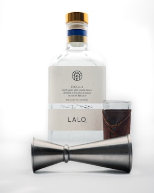 A bottle of tequila with a white label and gold cap is placed behind a metal jigger and a small glass with a brown leather sleeve.
