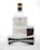 A bottle of tequila with a white label and gold cap is placed behind a metal jigger and a small glass with a brown leather sleeve.