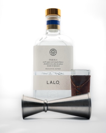 A bottle of tequila with a white label and gold cap is placed behind a metal jigger and a small glass with a brown leather sleeve.
