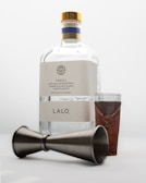 A bottle of tequila labeled 'LALO' with a wooden-capped top, accompanied by a shot glass wrapped in a leather holder and a metal jigger lying on its side.