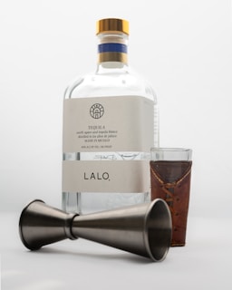 A bottle of tequila labeled 'LALO' with a wooden-capped top, accompanied by a shot glass wrapped in a leather holder and a metal jigger lying on its side.