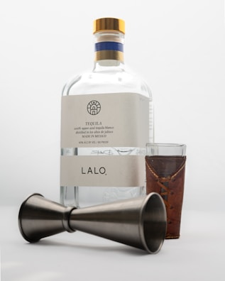 A bottle of tequila labeled 'LALO' with a wooden-capped top, accompanied by a shot glass wrapped in a leather holder and a metal jigger lying on its side.