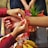 Two hands are tying a red thread bracelet around another person's wrist, suggesting a ceremonial or cultural practice. The scene includes a variety of items such as flowers and fruits, arranged on a table.