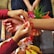 Two hands are tying a red thread bracelet around another person's wrist, suggesting a ceremonial or cultural practice. The scene includes a variety of items such as flowers and fruits, arranged on a table.