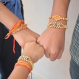 Three hands close together, each wearing a colorful, beaded bracelet. The bracelets feature intricate designs with red, orange, gold, and pearl-like beads. The hands are in a gesture that suggests unity or solidarity.