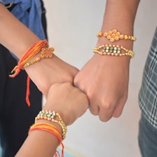 Three hands close together, each wearing a colorful, beaded bracelet. The bracelets feature intricate designs with red, orange, gold, and pearl-like beads. The hands are in a gesture that suggests unity or solidarity.