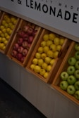 Wooden crates filled with fresh fruits, including yellow lemons, red apples, and green apples, are neatly arranged in a market display. The sign above references lemonade, suggesting a theme or focus on fresh produce.