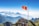 A large group of people stand on a viewing platform atop a mountain, with a red flag featuring a yellow star prominently displayed on a tall flagpole. The background consists of expansive mountain ranges covered in mist or clouds, under a clear blue sky.