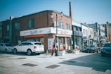 A street view of a city area with a mix of modern and traditional elements. The scene includes a row of buildings with signage in Korean, cars parked along the side, and a person walking on the sidewalk. Bright sunlight enhances the clear blue sky, creating distinct shadows on the ground.