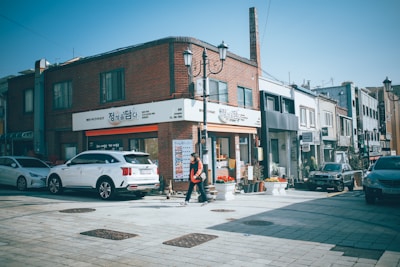 A street view of a city area with a mix of modern and traditional elements. The scene includes a row of buildings with signage in Korean, cars parked along the side, and a person walking on the sidewalk. Bright sunlight enhances the clear blue sky, creating distinct shadows on the ground.