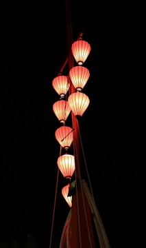 A series of red lanterns are arranged vertically against a dark background. The lanterns are glowing softly, illuminating their surroundings. The structure holding them appears to be part of a larger installation, possibly a decorative or festive display.