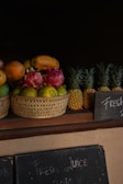 A collection of tropical fruits is displayed in wicker baskets. The assortment includes dragon fruits, papayas, and mangoes in one basket, while several whole pineapples are neatly arranged next to it. A dark background emphasizes the vibrant colors of the fruits. Below the baskets, there is a chalkboard sign with the words 'Fresh Juice' written in chalk.