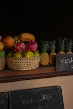A collection of tropical fruits is displayed in wicker baskets. The assortment includes dragon fruits, papayas, and mangoes in one basket, while several whole pineapples are neatly arranged next to it. A dark background emphasizes the vibrant colors of the fruits. Below the baskets, there is a chalkboard sign with the words 'Fresh Juice' written in chalk.