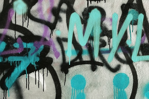 The image features graffiti art painted on a textured wall. Vibrant turquoise letters are prominently displayed, accompanied by black outlines and drips. Purple accents provide additional detail and depth to the artwork. The composition is abstract and freeform, conveying a sense of urban expression.