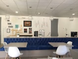 A modern interior space with a blue upholstered bench, white chairs, and tables. The wall features motivational quotes and words arranged in creative patterns. Decorative elements include hanging light fixtures and potted orchids. The overall ambiance is clean and inspiring.