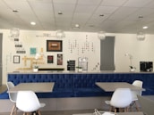 A modern interior space with a blue upholstered bench, white chairs, and tables. The wall features motivational quotes and words arranged in creative patterns. Decorative elements include hanging light fixtures and potted orchids. The overall ambiance is clean and inspiring.