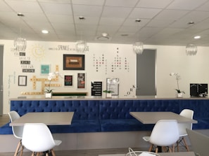 A modern interior space with a blue upholstered bench, white chairs, and tables. The wall features motivational quotes and words arranged in creative patterns. Decorative elements include hanging light fixtures and potted orchids. The overall ambiance is clean and inspiring.