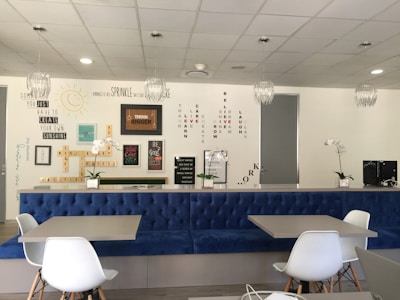 A modern interior space with a blue upholstered bench, white chairs, and tables. The wall features motivational quotes and words arranged in creative patterns. Decorative elements include hanging light fixtures and potted orchids. The overall ambiance is clean and inspiring.