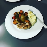 A white plate holds a serving of grilled chicken, various roasted vegetables, and a portion of scrambled eggs. The vegetables appear to include zucchini, carrots, and red peppers, all cooked to a golden brown.