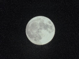 A full moon is prominently displayed against a dark, starless sky. The moon's surface features are clearly visible, including various craters and shading that give it a textured appearance.