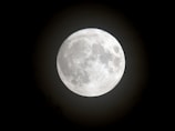 A large, bright full moon with detailed surface features such as craters and lunar maria, set against a dark night sky.