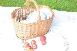 A collapsible linen picnic stool set up on a grassy lawn beside a woven picnic basket.