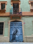 A green building facade with orange trim features a balcony above a large blue double door. The door has artwork depicting a black and white bird with intricate details, set in a circular design. The windows on either side have ornate frames, and an open balcony door reveals part of the interior.