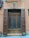 A wooden door with two side panels and a transom is set within a textured wall. The door features frosted glass and is framed by shutters. An arrangement of cables and electrical boxes is mounted on the wall to the left. The building facade is painted in earthy tones.