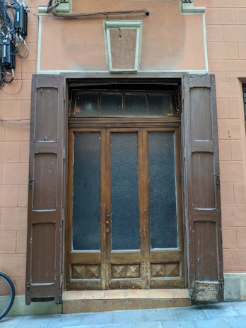 A wooden door with two side panels and a transom is set within a textured wall. The door features frosted glass and is framed by shutters. An arrangement of cables and electrical boxes is mounted on the wall to the left. The building facade is painted in earthy tones.