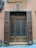 A wooden door with two side panels and a transom is set within a textured wall. The door features frosted glass and is framed by shutters. An arrangement of cables and electrical boxes is mounted on the wall to the left. The building facade is painted in earthy tones.