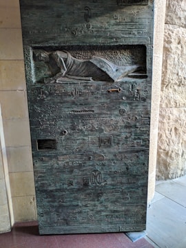 A textured metal door featuring intricate carvings and engravings, including a relief of a reclining figure in medieval garb near the top. The surface is adorned with various abstract symbols and geometric shapes, providing a sense of mystery and historical significance.