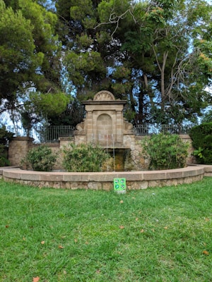 A stone fountain set against a backdrop of dense green trees and bushes. The fountain features a central arched section with water spouting from underneath into a stone basin surrounded by manicured shrubbery. In front of the fountain is a neatly trimmed grassy area, with a vibrant green and blue sign on the grass.