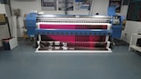 A large digital printing machine inside a room, producing a vivid banner with pink, black, and white colors. The machine has a blue exterior with multiple control buttons and switches, situated against a neutral-colored wall with a few posters and signs.