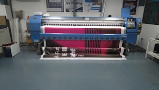A large digital printing machine inside a room, producing a vivid banner with pink, black, and white colors. The machine has a blue exterior with multiple control buttons and switches, situated against a neutral-colored wall with a few posters and signs.