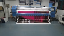 A large digital printing machine inside a room, producing a vivid banner with pink, black, and white colors. The machine has a blue exterior with multiple control buttons and switches, situated against a neutral-colored wall with a few posters and signs.