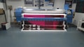 A large digital printing machine inside a room, producing a vivid banner with pink, black, and white colors. The machine has a blue exterior with multiple control buttons and switches, situated against a neutral-colored wall with a few posters and signs.