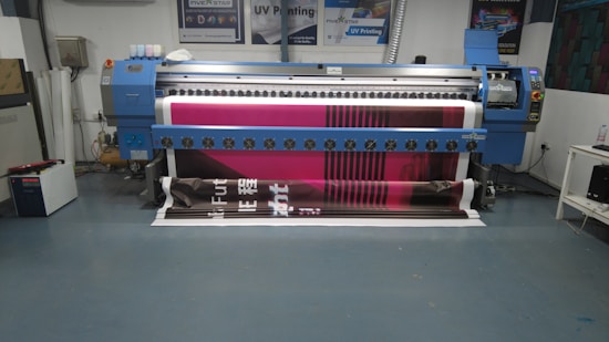 A large digital printing machine inside a room, producing a vivid banner with pink, black, and white colors. The machine has a blue exterior with multiple control buttons and switches, situated against a neutral-colored wall with a few posters and signs.