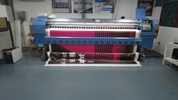 A large digital printing machine inside a room, producing a vivid banner with pink, black, and white colors. The machine has a blue exterior with multiple control buttons and switches, situated against a neutral-colored wall with a few posters and signs.